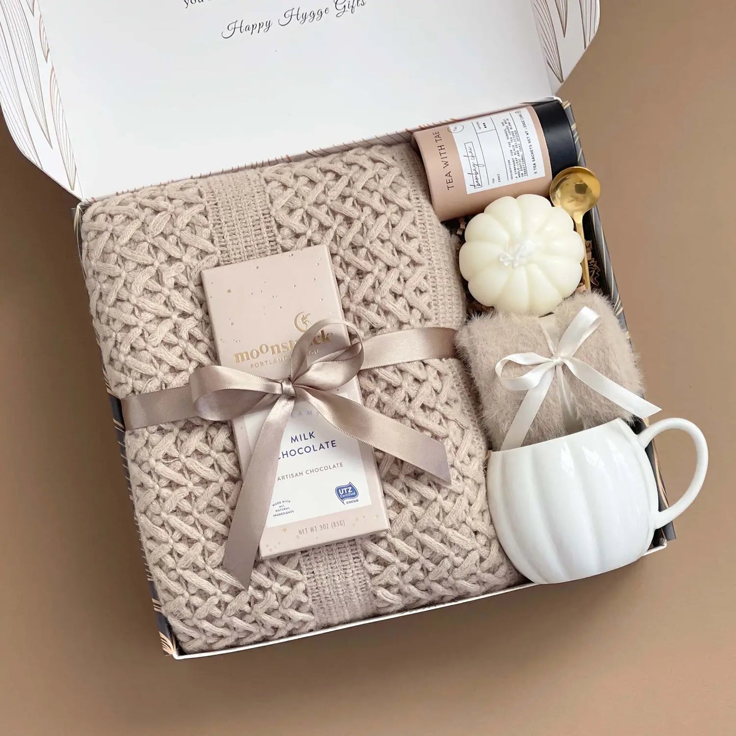 Gift box with a knitted blanket, mug, and small items on a beige background


